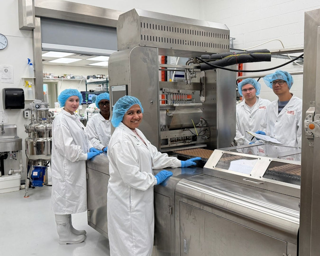 Two GNL staff in lab coats, hairnets and gloves working beside gummy moulds and a stainless mixing vessel on the Edmonton production floor