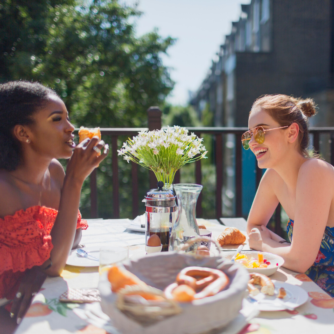 two friends sit outside on a balcony on a sunny day enjoying brunch